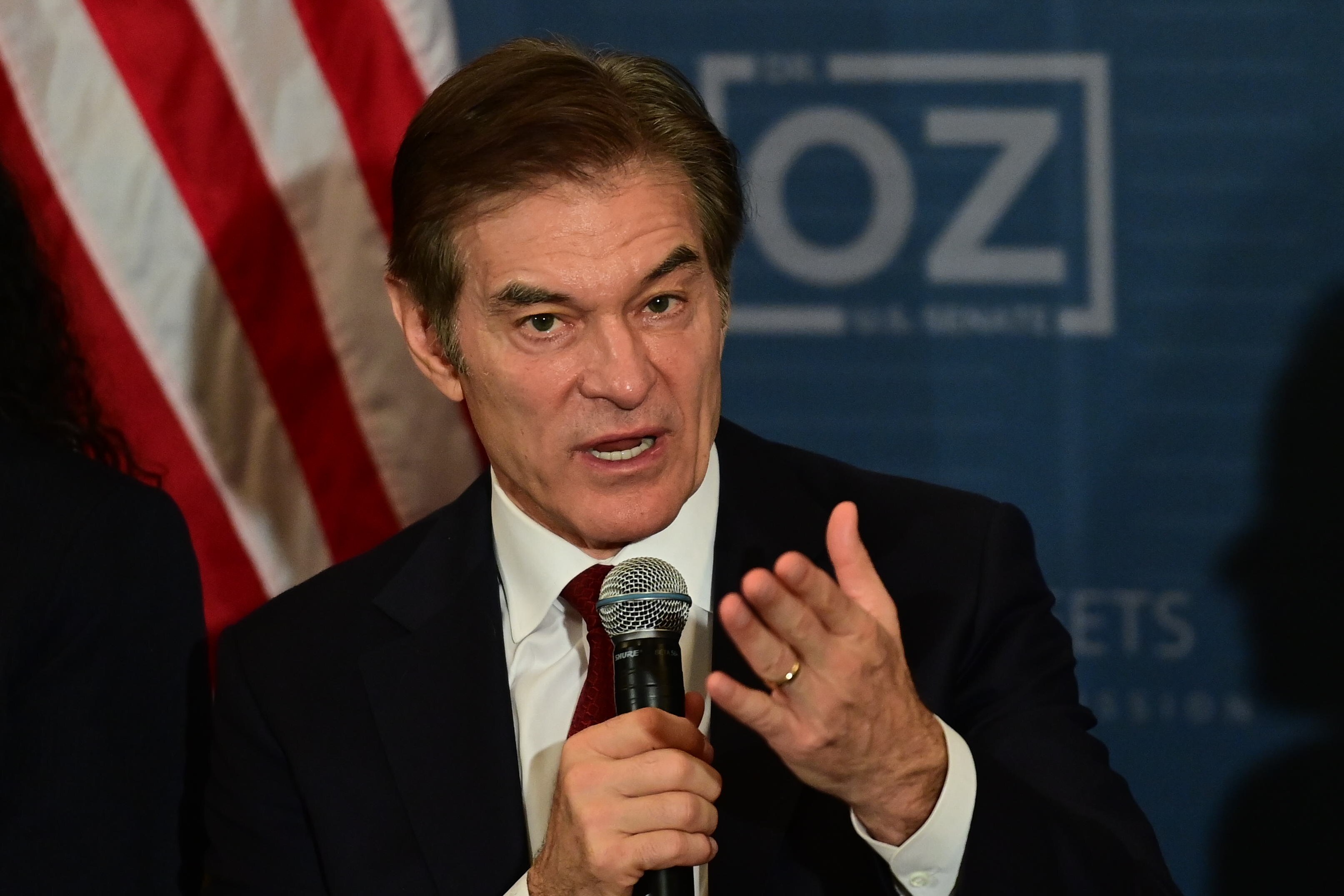 Mehmet Oz speaks into a microphone while gesturing with his left hand. Oz is wearing a dark suit and is in front of an American flag.