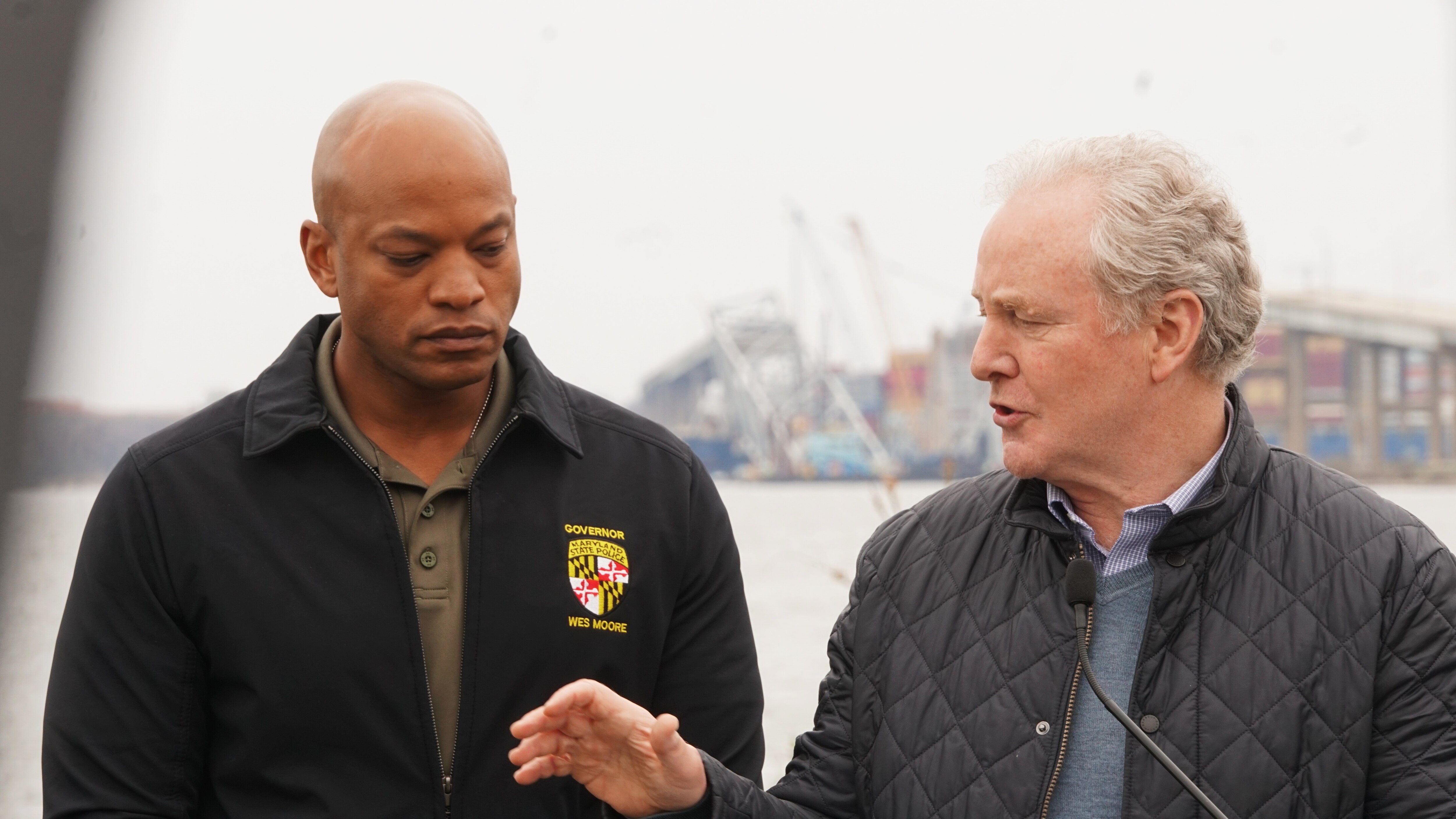 Two men stand next to each other and one speaks into a podium microphone.