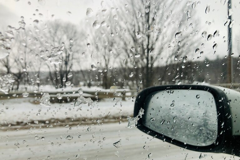 A picture of a vehicle window from the perspective of someone inside the vehicle, looking out. The window is covered in rain and melting frost, with a rearview mirror in the photo and an icy landscape beyond.
